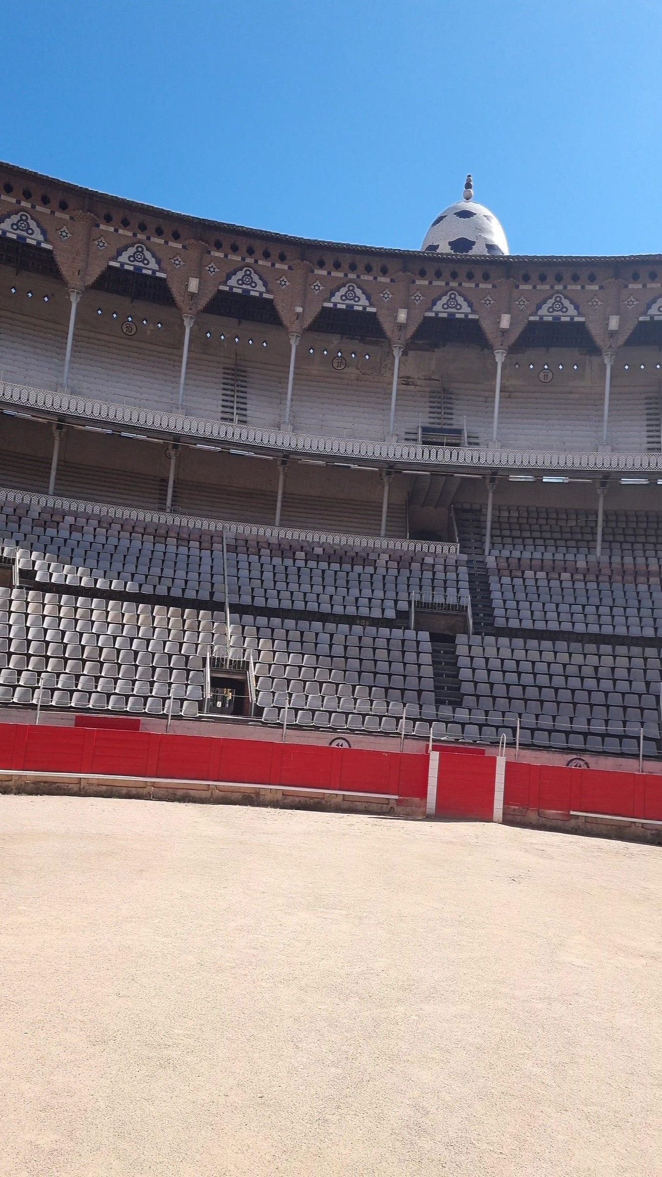 An empty bullring with rows of grey seats, a red barrier and a sandy floor.